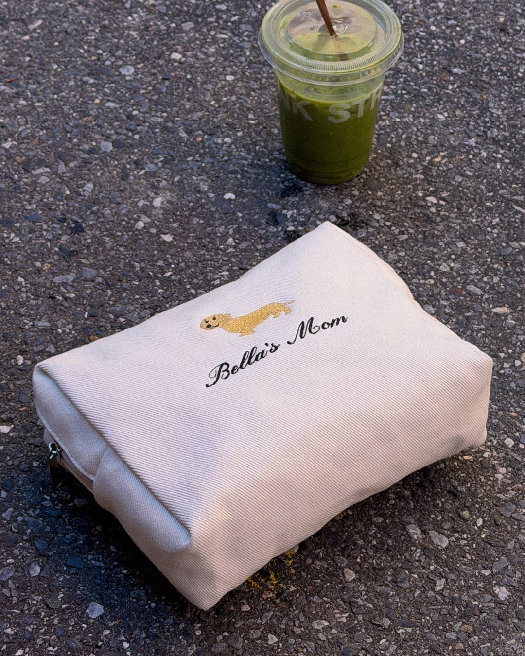 White pouch with 'Bella's Mom' embroidery on a concrete surface next to matcha