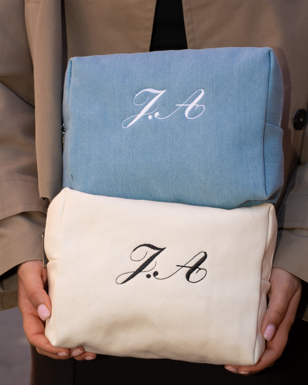 Two embroidered pouches, one blue and one beige, held by hands with 'J.A' monogrammed on them.