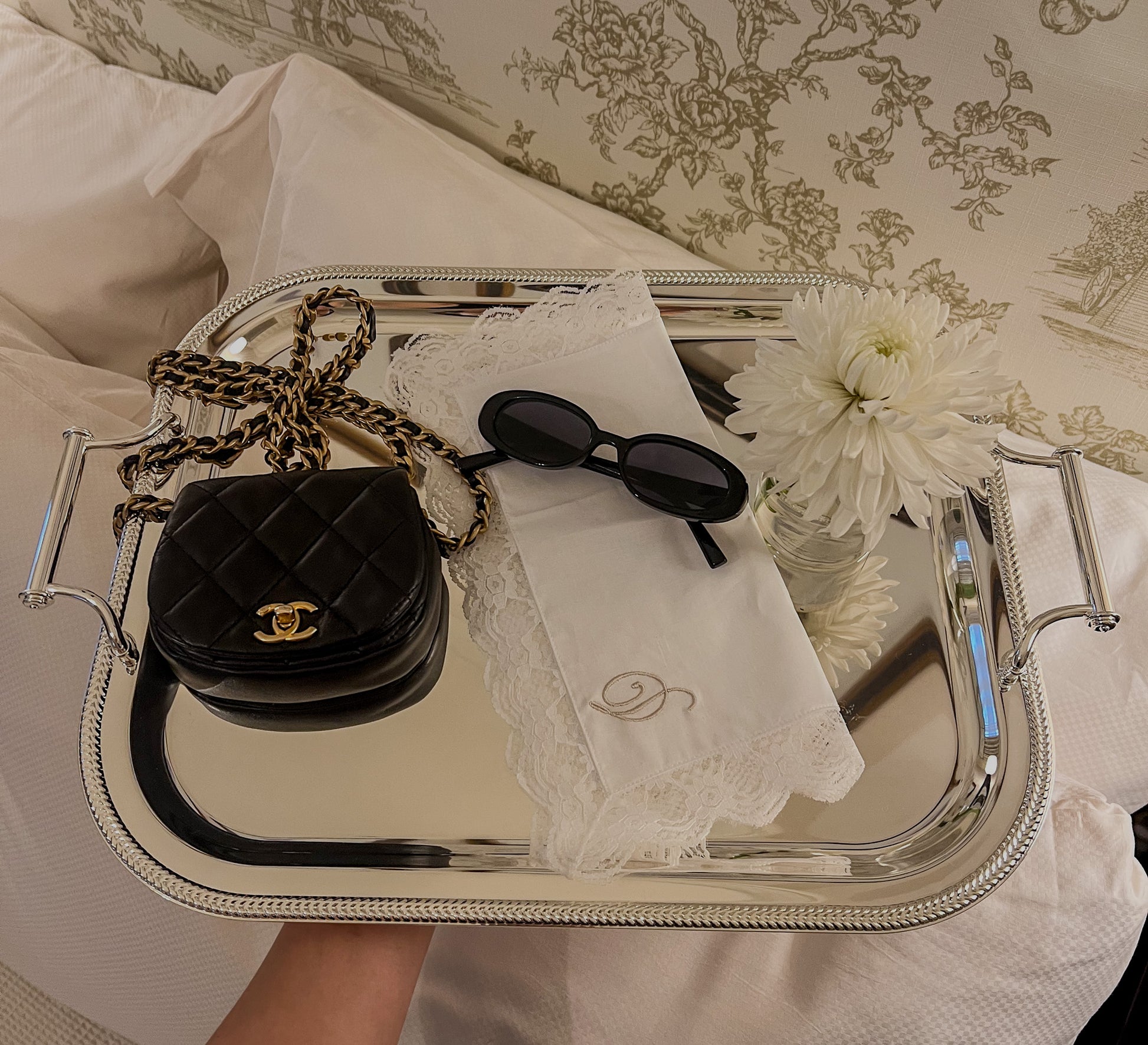 Black handbag, sunglasses, and white flowers on a silver tray with floral wallpaper background