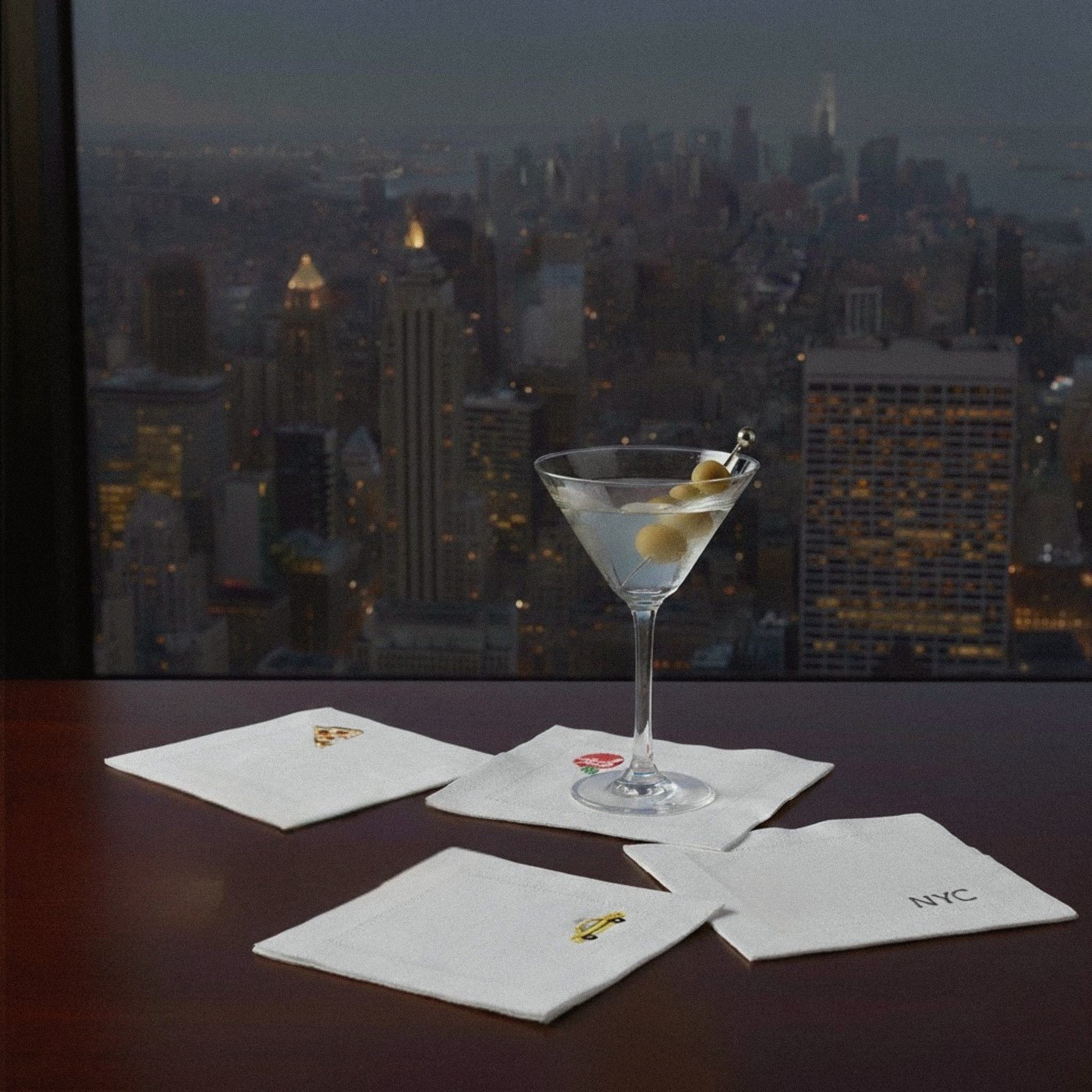 Martini glass with olives on a table in front of a city skyline at night.